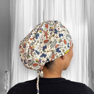 May include: A patterned surgical scrub cap featuring a cream background with colorful illustrations of crawfish, musical instruments, and other New Orleans-themed designs. The cap has a tie in the back and a yellow accent.