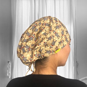 May include: A beige scrub cap featuring a repeating pattern of gray sock monkey figures with red and white accents. The cap has a drawstring closure and a small yellow tag.