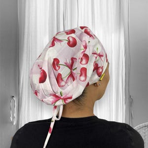May include: A white and pink patterned surgical scrub cap featuring a cherry and bow design. The cap is tied at the back with matching fabric ties. The background is a white curtain.