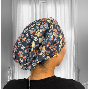 Puede incluir: Un gorro de quirófano azul con un estampado de huellas de patas de colores. El gorro tiene un lazo en la parte posterior.