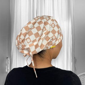 May include: A brown and white checkered scrub hat with a pattern of smiley faces. The hat has a tie at the back.