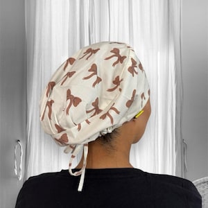 Puede incluir: Un gorro quirúrgico blanco con un patrón repetido de lazos marrones. El gorro se ata en la parte posterior con lazos blancos. El gorro lo lleva una persona que lleva una camisa negra.