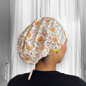 May include: A patterned scrub cap featuring a repeating design of suns, flowers, ice cream cones, and beach balls in orange, yellow, pink, and blue on a cream background. The cap has a tie in the back.