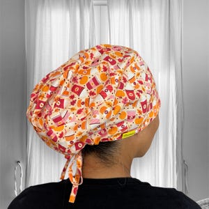 May include: A white scrub cap with a colorful pattern of orange, red, and brown. The pattern features leaves, pumpkins, and coffee cups. The cap has a tie at the back.