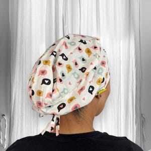May include: A white scrub cap featuring a pattern of speech bubbles in pink, yellow, blue, and black. The cap is tied at the back with fabric ties. The background is a white curtain.