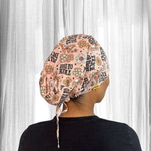 May include: A pink patterned scrub cap featuring flowers, butterflies, and the words "Trick or Treat". The cap is tied at the back and is worn by a person wearing a black shirt. The background is a white curtain.