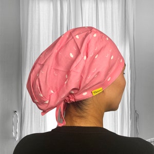 Puede incluir: Un gorro de quirófano rosa con un estampado blanco de ojos y labios. El gorro tiene un lazo en la parte posterior y una etiqueta amarilla que dice "Tamer".