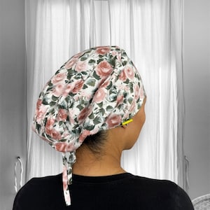 May include: A white scrub cap with a pink and green floral pattern featuring roses. The cap has a long tie that can be tied at the back of the neck.