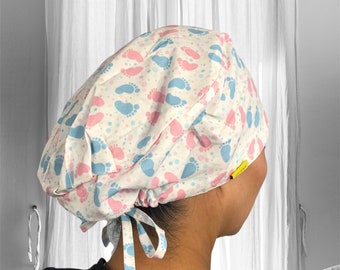 Puede incluir: Un gorro de quirófano blanco con un patrón de huellas de pies de bebé azul y rosa. El gorro tiene un lazo en la parte posterior.