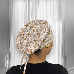Puede incluir: Un gorro quirúrgico estampado con un diseño floral y de lazo en tonos rosa, marrón y blanco. El gorro tiene lazos en la parte posterior. El diseño incluye las palabras "fuerza", "esperanza" y "coraje".
