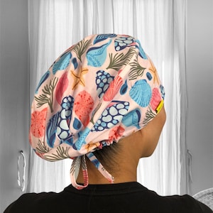 Puede incluir: Un gorro quirúrgico rosa claro con un estampado de conchas marinas de colores. El diseño incluye varias conchas, estrellas de mar y algas en tonos azules, coral y verde. El gorro tiene un cierre de lazo en la parte posterior.