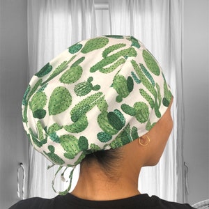 May include: A white scrub cap with a green cactus print. The cap has a tie at the back.