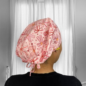 Puede incluir: Un gorro quirúrgico rosa con un patrón de lazos y estrellas rosas. El gorro tiene un lazo en la parte posterior.