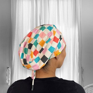 Peut inclure: Un bonnet chirurgical à motifs avec un motif de damier répétitif. Le bonnet présente des carrés roses, oranges, verts, noirs et bleus. Il est noué dans le dos avec une ficelle blanche.