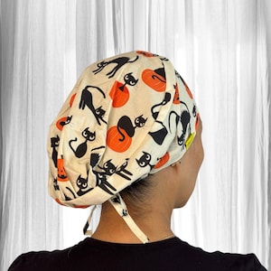 May include: A scrub cap featuring a cream-colored background with a repeating pattern of black cats and orange pumpkins. The fabric head covering has a gathered back design with tie closures at the nape of the neck and a small yellow brand tag.