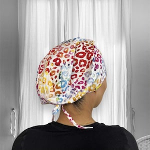 May include: A colorful, patterned surgical scrub cap. The cap features a white background with a vibrant leopard print design in shades of red, yellow, blue, and purple. The cap is tied at the back with matching fabric ties.