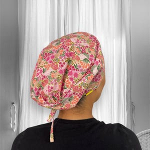 May include: A floral patterned scrub cap in shades of pink, orange, and green. The cap is tied at the back and is worn by a person. The background is a white curtain.