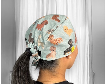 May include: A blue and grey surgical scrub hat with a pattern of dachshunds. The hat has a tie at the back and is worn by a person with long brown hair.