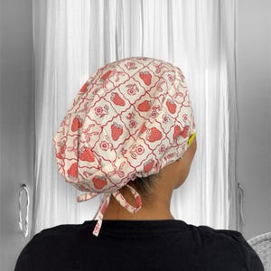 Puede incluir: Un gorro quirúrgico blanco con un estampado repetido de fresas, flores y lazos rojos. El gorro se ata en la parte posterior con lazos de tela a juego. El fondo muestra una vista borrosa de una ventana y cortinas.