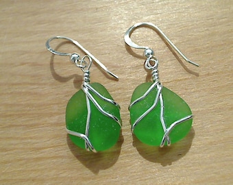May include: A pair of handmade earrings featuring smooth, green sea glass stones wrapped in silver-colored wire. The earrings have a simple hook design for pierced ears, showcasing a natural, beachy aesthetic.