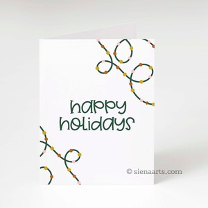 May include: A white card with green text that says "happy holidays". The card is decorated with a green string of lights with pink, orange, and gold bulbs.