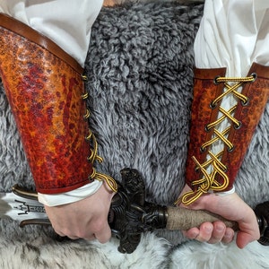 May include: A pair of brown and orange leather arm guards with yellow lacing. The arm guards are worn over white sleeves and held by a person holding a sword with a detailed handle. The leather has a textured, mottled appearance.