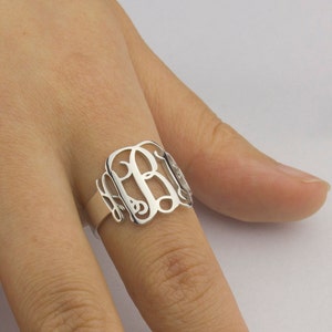May include: A silver ring with a monogram design. The ring is worn on a finger.