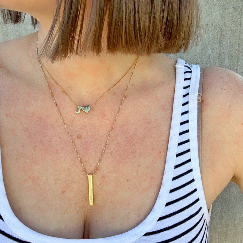 May include: A woman wearing a white and black striped tank top with two gold necklaces. One necklace has a heart pendant with the letter J on it. The other necklace has a long, thin, rectangular pendant.