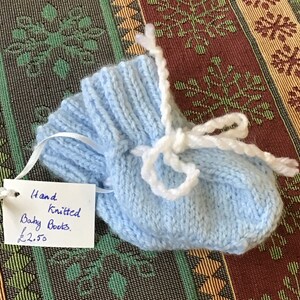 May include: A pair of light blue hand-knitted baby booties with white ties. The booties are on a patterned fabric background. The price tag reads "Hand Knitted Baby Boots £2.50".