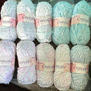 May include: Ten skeins of Peter Pan Cupcake yarn arranged in two rows. The yarn features various pastel colour combinations including light blue, white, and pink variegated strands. Each skein has a white paper label with the text Cupcake.