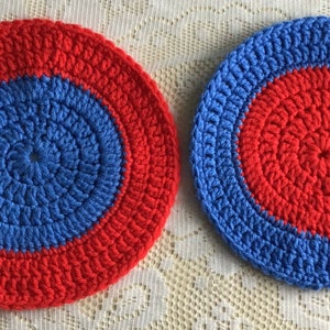 May include: Two crocheted round coasters with a concentric circle design. The larger coaster is red with a blue centre circle. The smaller coaster is blue with a red centre circle.