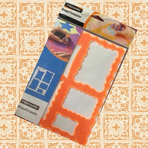 May include: A set of four orange plastic shape templates for crafting. The templates are in the shape of frames with different decorative edges. The packaging includes the text "Fiskars ShapeTemplate".