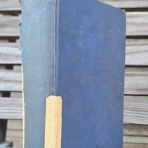 May include: A blue hardcover book with the title "The Mill on the Floss" written in cursive on the front cover. The book has a yellowed label on the spine with the letters "S, Y, W, D, S" written on it. The book is sitting on a wooden surface.