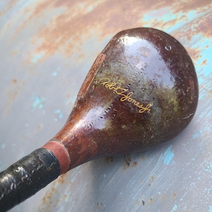 May include: Vintage wooden golf club with a dark brown head and a black grip. The club head has the inscription "Robert T. Jones Jr." in gold lettering. The club shows signs of wear, suggesting it is an antique.
