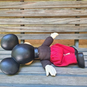 May include: A plush Mickey Mouse doll lying on a wooden bench. The doll is wearing a red skirt and has black ears and a black face.