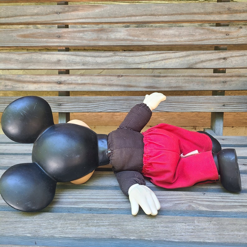 May include: A plush Mickey Mouse doll lying on a wooden bench. The doll is wearing a red skirt and has black ears and a black face.