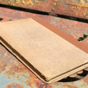 May include: A vintage book with a brown spine and a tan cover. The book is lying on a rusty metal surface.