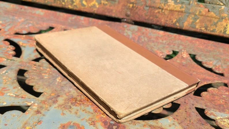 May include: A vintage book with a brown spine and a tan cover. The book is lying on a rusty metal surface.