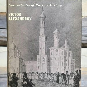May include: A vintage book cover with a black and white illustration of the Kremlin in Moscow, Russia. The title of the book is "The Kremlin" and the subtitle is "Nerve-Centre of Russian History". The author is Victor Alexandrov.