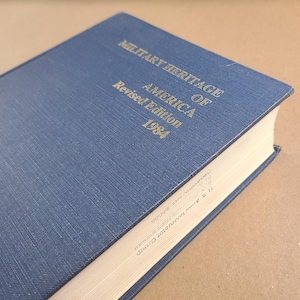 May include: A blue hardcover book titled "MILITARY HERITAGE OF AMERICA" with gold lettering. The book is a revised edition from 1984. The book's cover has a textured appearance. The book's pages are white.