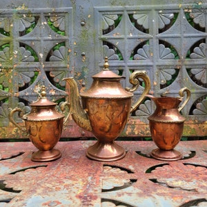 May include: Three antique copper teapots with ornate brass accents. The largest teapot has a spout and a handle on the side. The smallest teapot has a lid and a handle on the side. The middle teapot has a lid and a handle on the side. The teapots are sitting on a rusty metal surface.