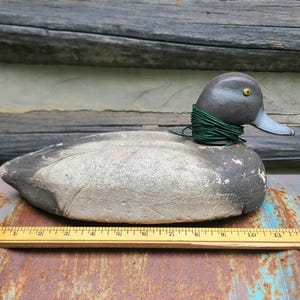 May include: A grey and white painted wooden duck decoy with a green string around its neck. The decoy has a yellow eye and is sitting on a rusty metal surface.
