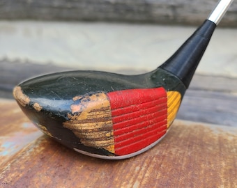 May include: Close-up of a vintage golf club head. The club head is black with red and yellow accents on the bottom. The club shows signs of wear and tear, with chipped paint and a wooden appearance.