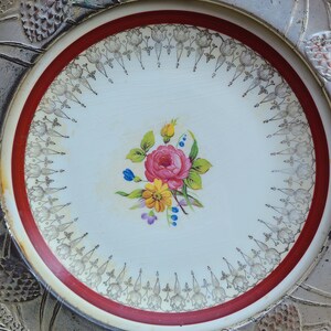 May include: A white ceramic plate with a red rim and a gold floral design. The center of the plate features a pink rose with yellow and blue flowers.
