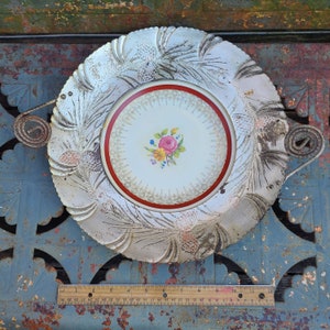 May include: A white ceramic serving dish with a raised, embossed design of pine needles. The dish has a central, round, white porcelain plate with a red rim and a floral design in the center. The dish has a metal handle on each side.