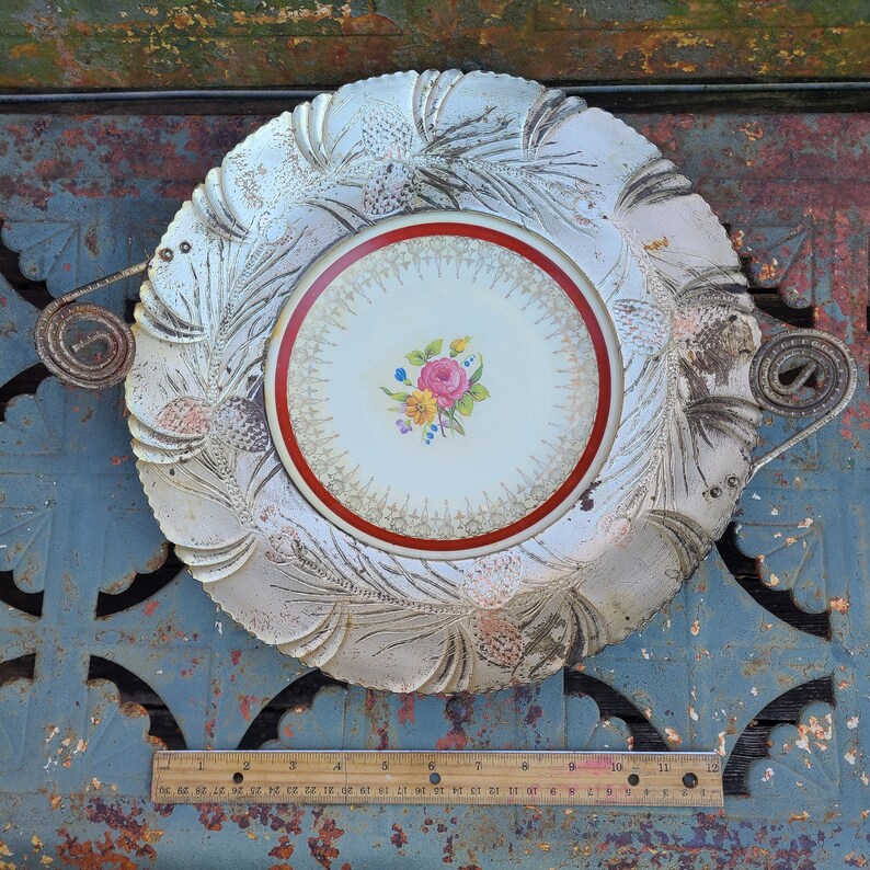May include: A white ceramic serving dish with a raised, embossed design of pine needles. The dish has a central, round, white porcelain plate with a red rim and a floral design in the center. The dish has a metal handle on each side.