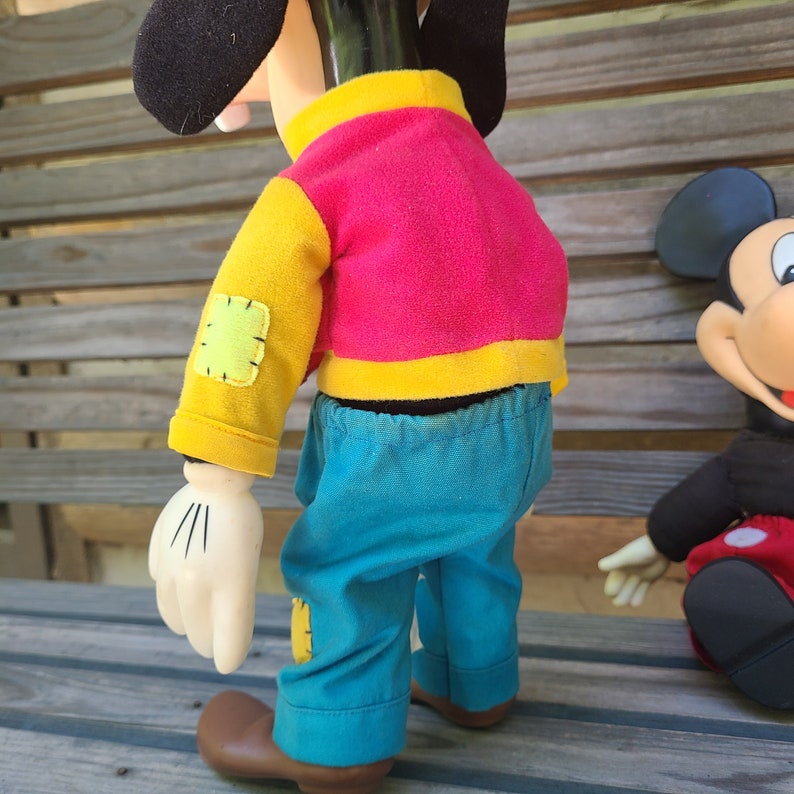 May include: A plush Goofy doll wearing a red and yellow jacket and blue trousers. The doll has a stitched patch on its arm and is standing on a wooden surface.