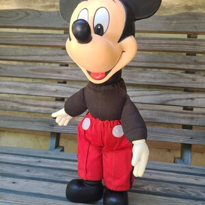 May include: A vintage plush Mickey Mouse toy with black ears, a red and white outfit, and a black and brown shirt. The toy is sitting on a wooden bench.