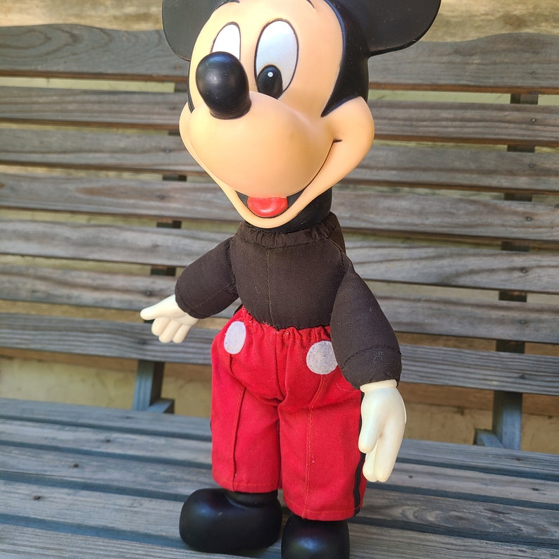 May include: A vintage plush Mickey Mouse toy with black ears, a red and white outfit, and a black and brown shirt. The toy is sitting on a wooden bench.