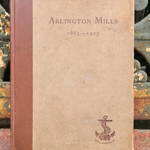May include: A brown book with a tan cover and a brown spine. The title is "Arlington Mills 1865-1925" and there is an anchor with the text "Skill Wins Favor" below it.